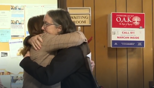 Photo of two people hugging in front of newly-installed Overdose Aid Kit. The two people are Sue Fischer and Jessica Geschke.